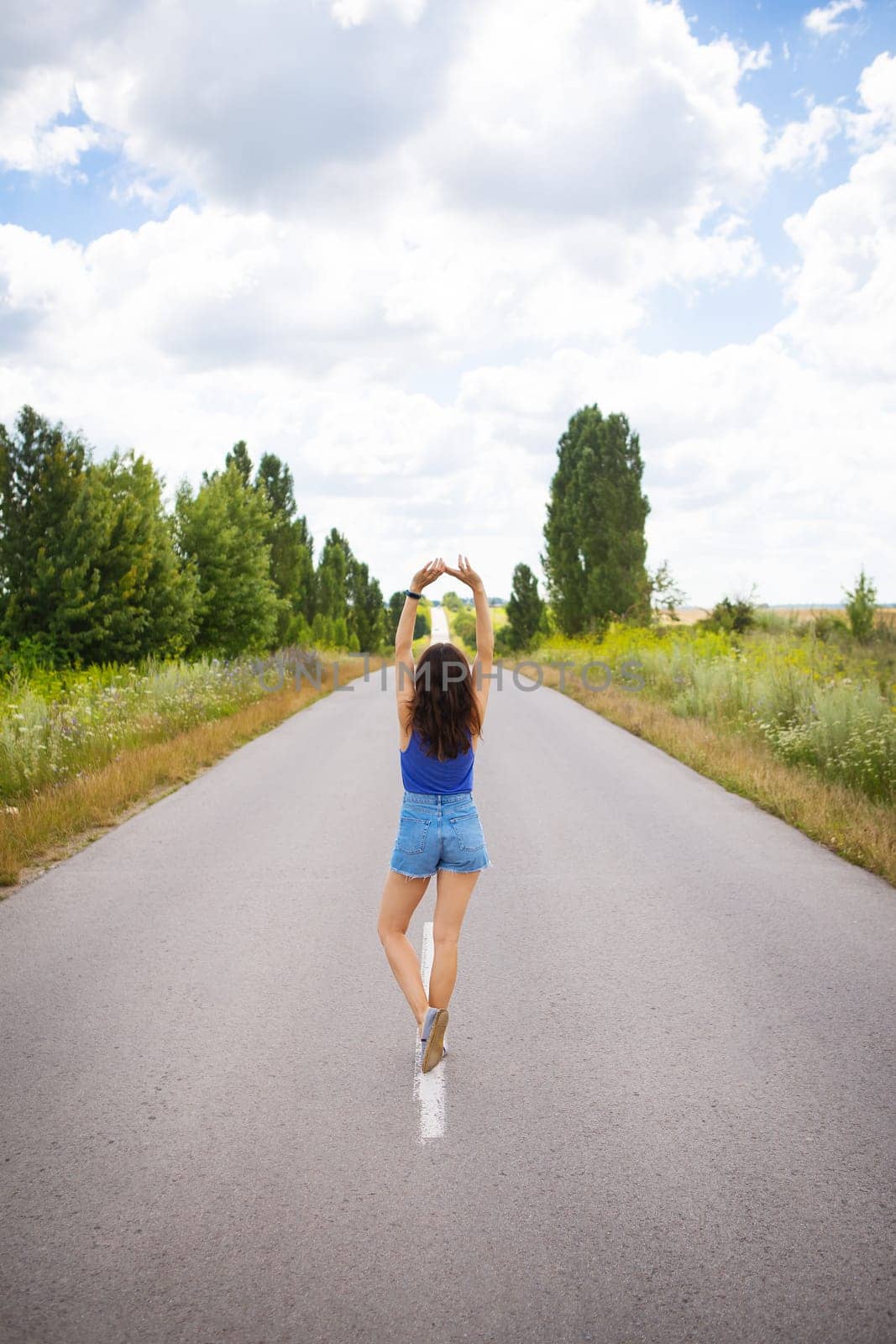 Attractive girl, stands posing, hands up in the middle of an asphalt road near the field, copies space for content