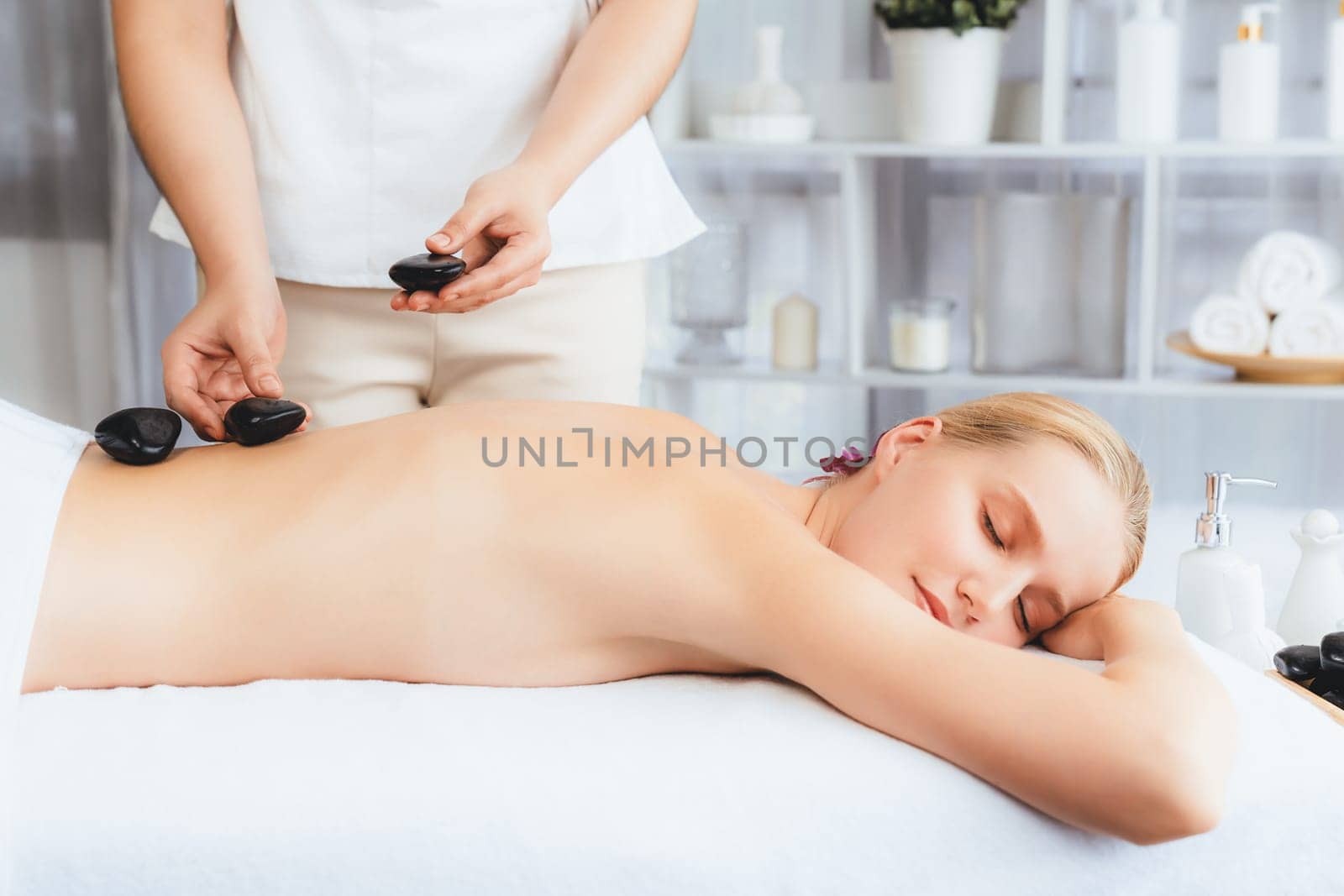 Hot stone massage at spa salon in luxury resort with day light serenity ambient, blissful woman customer enjoying spa basalt stone massage glide over body with soothing warmth. Quiescent