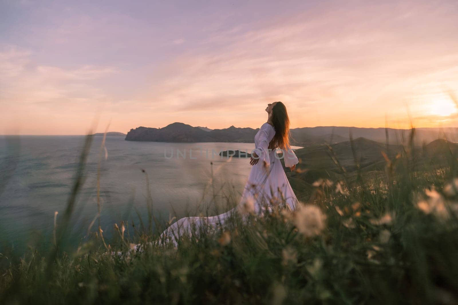 A woman in a white dress stands on a grassy hill overlooking the ocean. The sky is a mix of orange and purple hues, creating a serene and peaceful atmosphere. The woman is enjoying the view