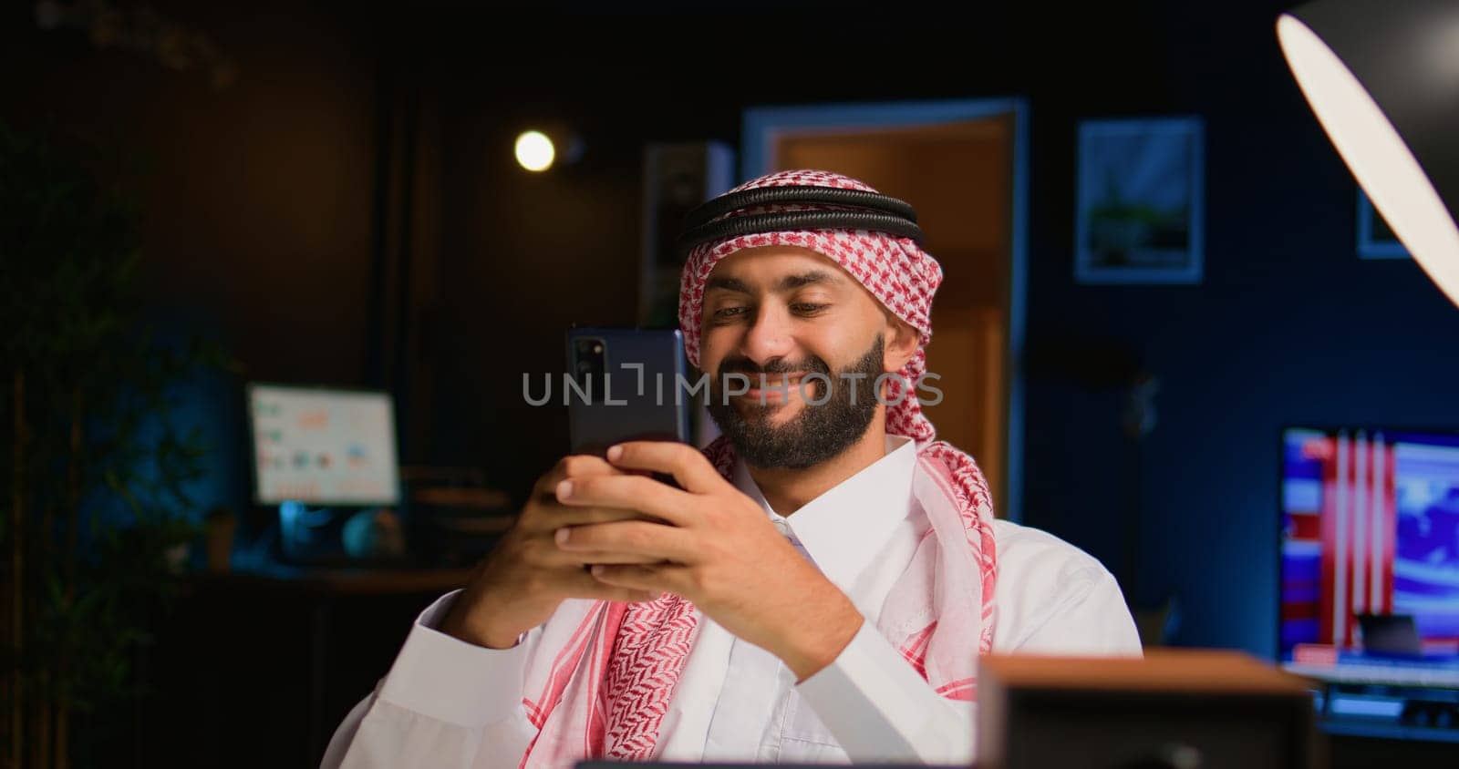 Smiling arab man at home in living room typing messages on smartphone. Middle Eastern person holding cellphone, enjoying relaxing leisure time talking with friends over online messaging app