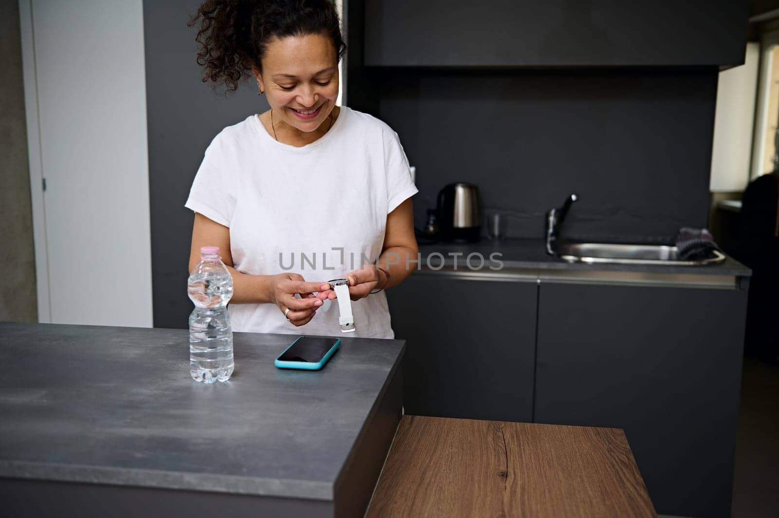 Smiling pretty woman in white t-shirt holding modern smart wrist watch with black mockup digital screen, standing at kitchen counter in the minimalist home interior. Copy space for advertising text.