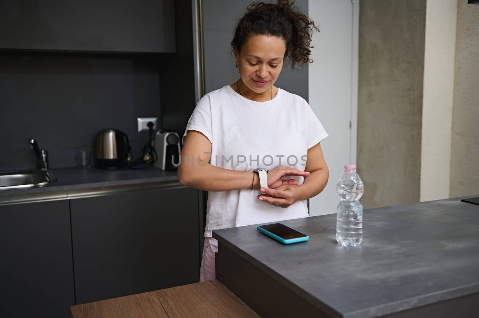 Smiling a pretty woman in white t-shirt putting on, wearing smart wrist watch with black mockup digital screen, standing at kitchen counter in the minimalist home interior. Copy advertising space.