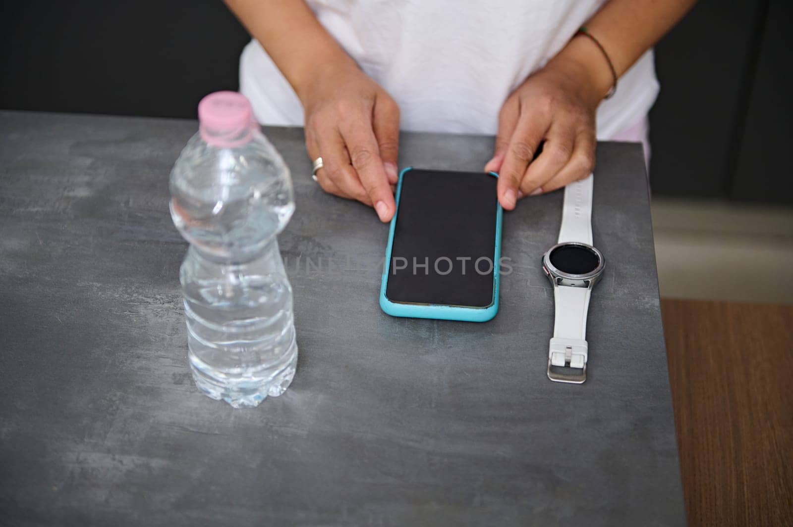 Cropped view hands of a woman standing at gray table with a smart watch, mobile phone and bottle of pure water. Blank black mockup touch screen with free space for advertising text.