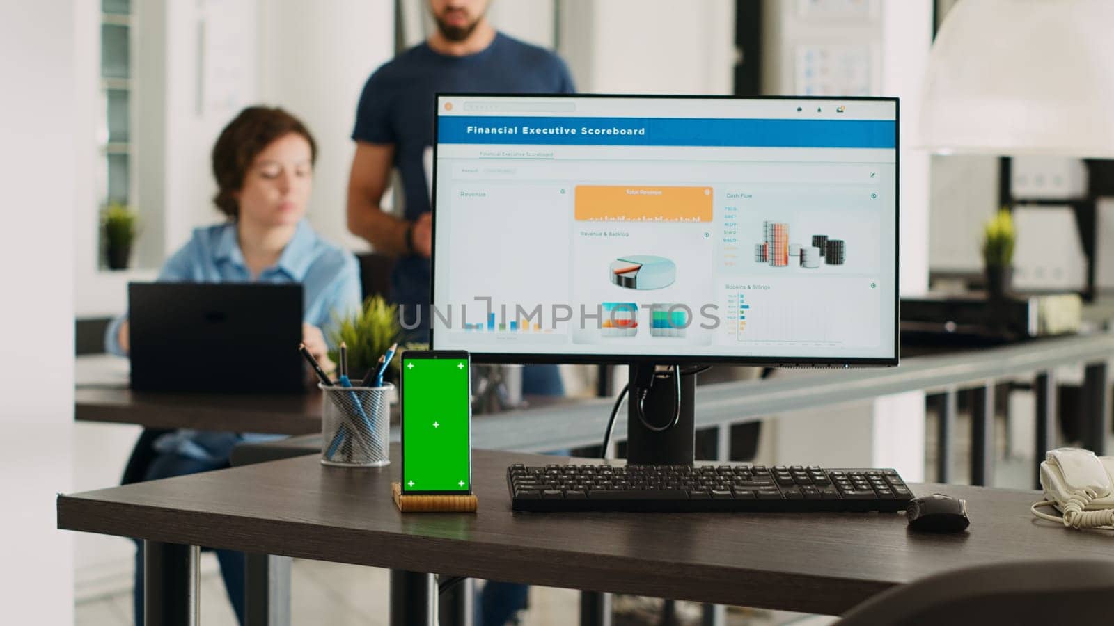 Persons conducting business tasks at agency workspace with cellphone displaying greenscreen at empty desk. Isolated copyspace mockup layout appears on workstation with mobile device.