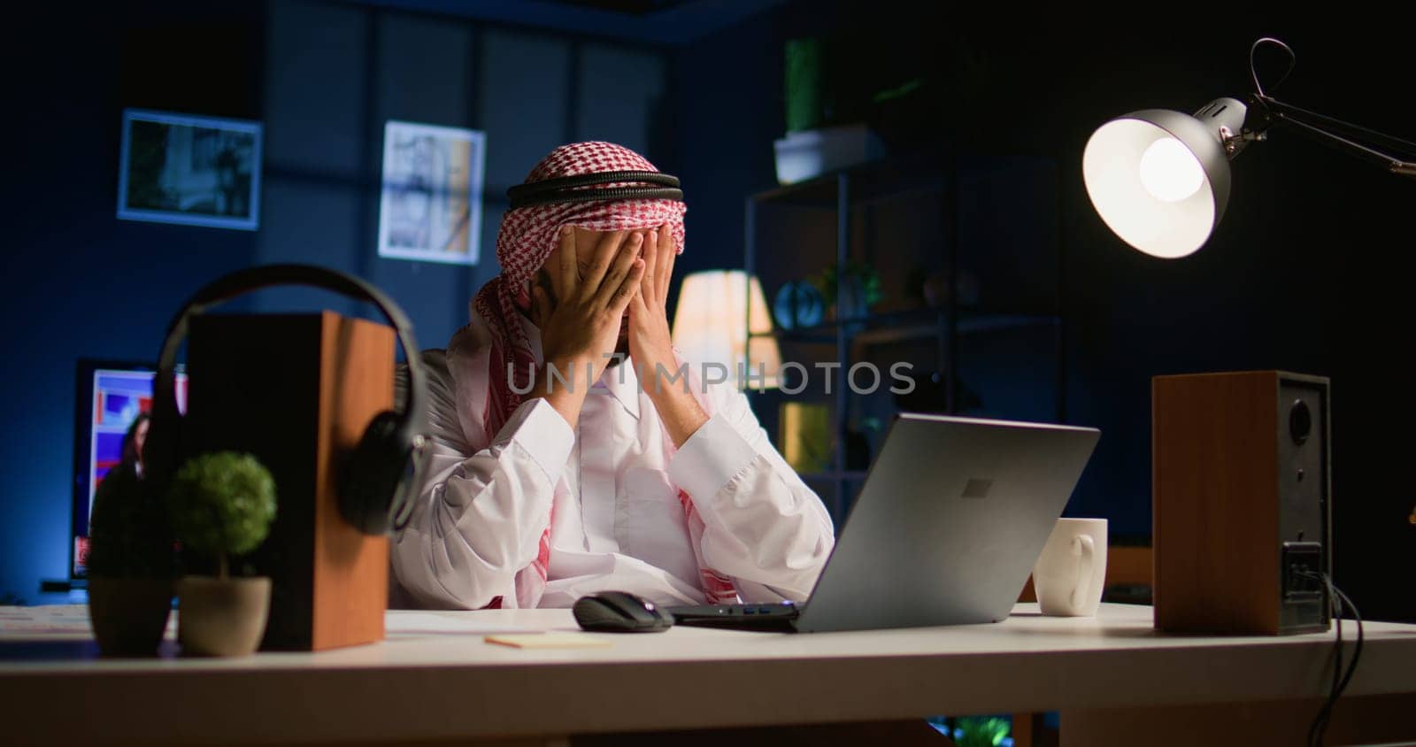 Tired arab businessman in traditional attire suffering migraine working at home office desk. Stressed Middle Eastern worker in living room answering emails getting headache from approaching deadline