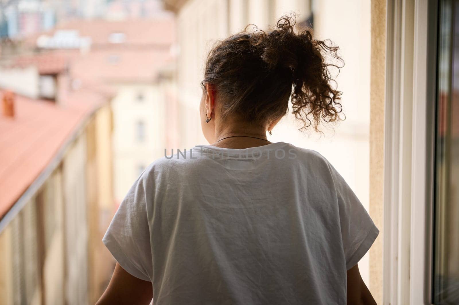 Rear view. Young woman in white t-shirt, enjoying the holidays morning, contemplating through the window the beautiful touristic city of Como with red roof tiles of cozy houses. People. Lifestyle