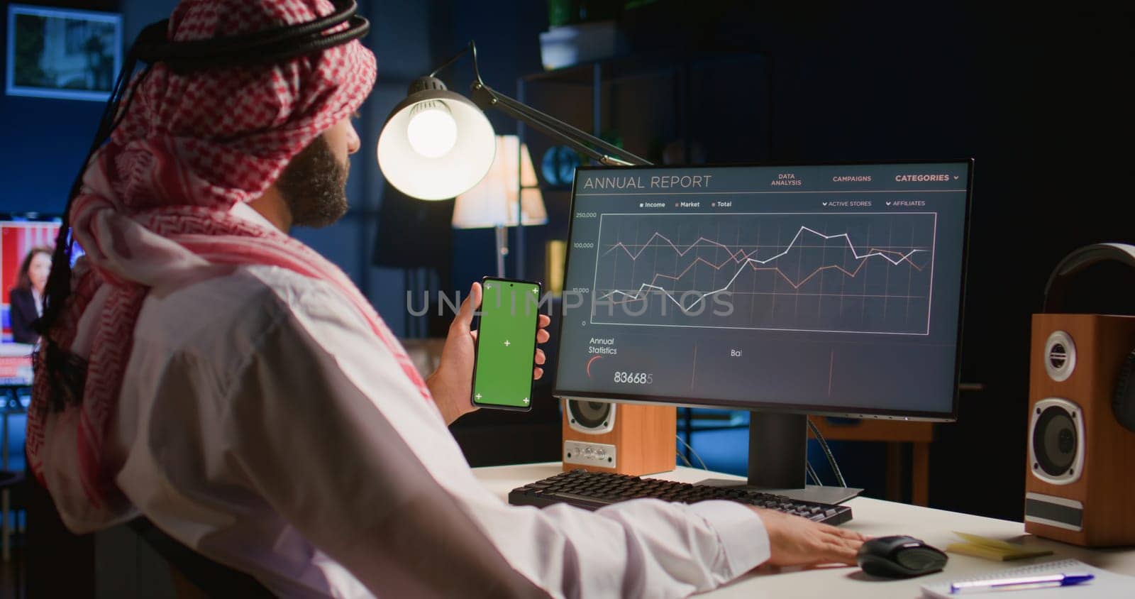 Man holding green screen smartphone while looking at business annual report graphs on computer. Self employed person with chroma key phone in hand performing tasks on device