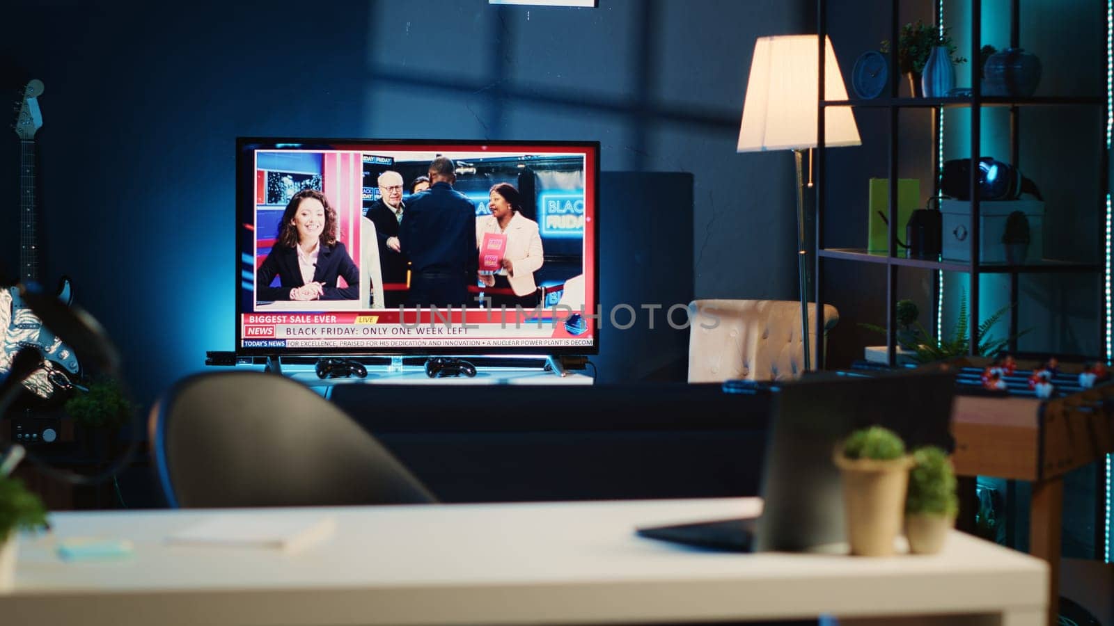 Apartment illuminated by RGB lights with TV broadcast showing Black Friday news story. Blurry foreground of work desk with laptop and mini house plants in dimly lit empty home studio interior