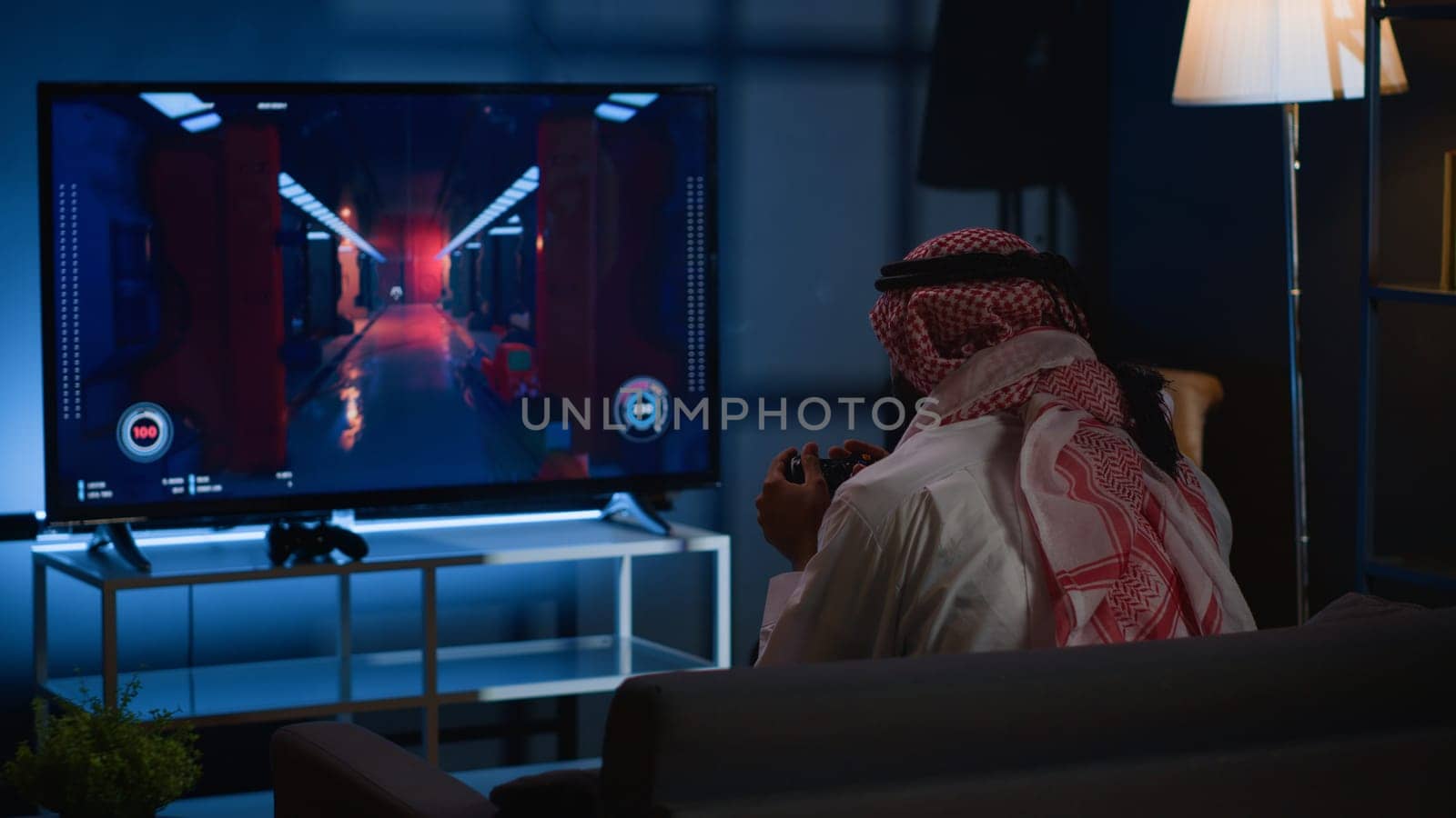 Arab man playing videogames on TV, relaxing after long day at work. Competitive gamer having fun on gaming console in stylish apartment living room, enjoying science fiction shooter game