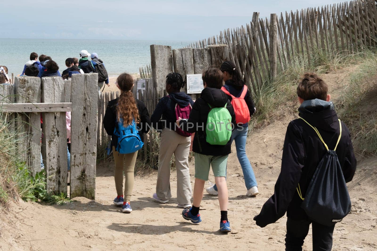 Utah Beach in Normandy, France. Unrecognizable children passing through wood sea fence, grass and sand dunes. Sunny sky light ble clouds and blue ocean in background. High quality photo