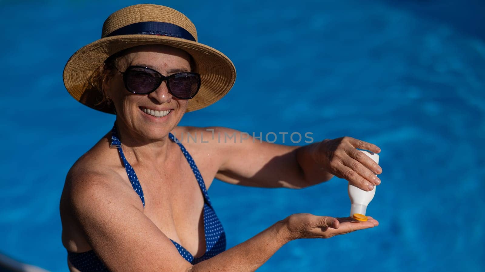 Portrait of an old woman in a straw hat, sunglasses and a swimsuit applying sunscreen to her skin while relaxing by the pool. by mrwed54