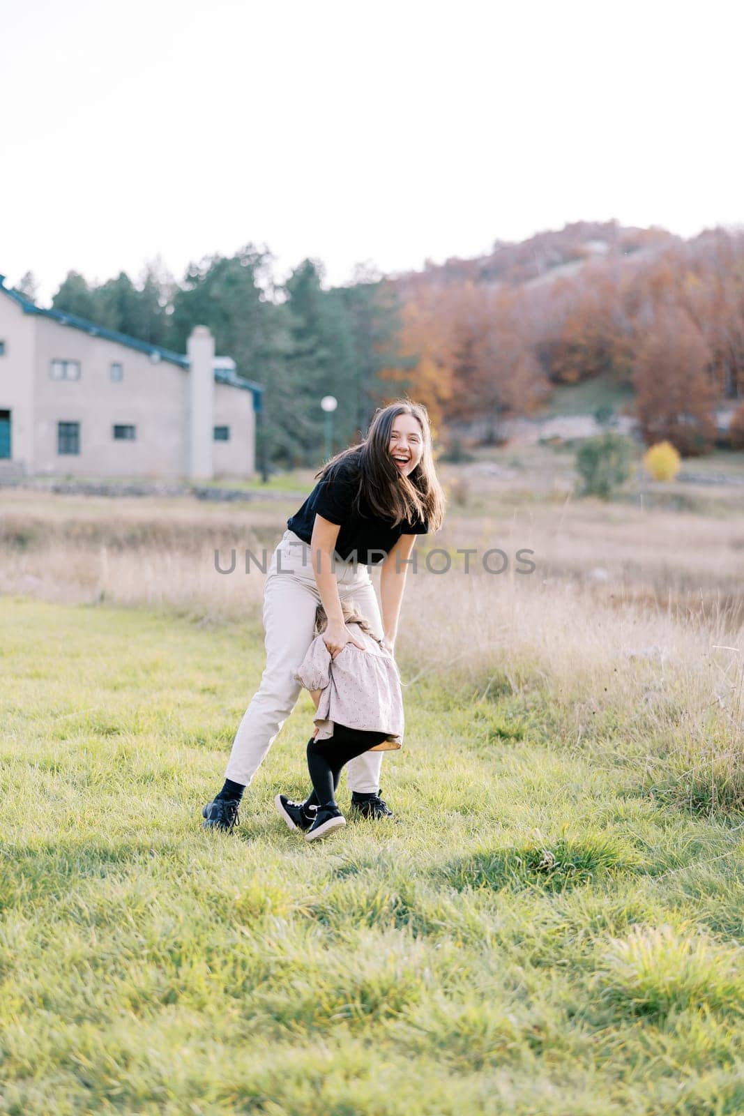 Laughing mother hugs little girl cuddled on her lap on green lawn. High quality photo