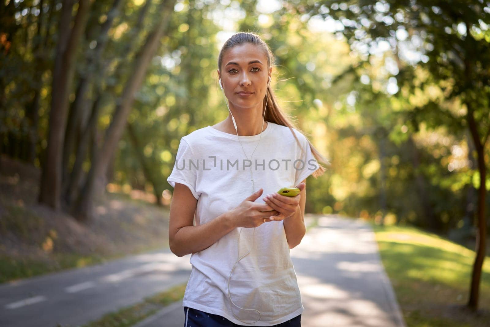 Portrait of confident female jogger listening to music through earphones while holding smartphone. Beautiful and fit woman runner in white shirt looking at camera, standing on road in park.