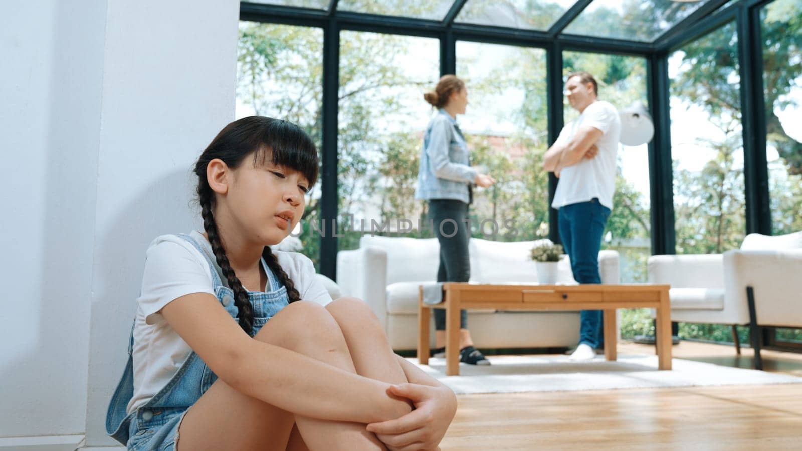 Stressed and unhappy young girl huddle in corner, cover her ears blocking sound of her parent arguing in background. Domestic violence at home and traumatic childhood develop to depression. Synchronos