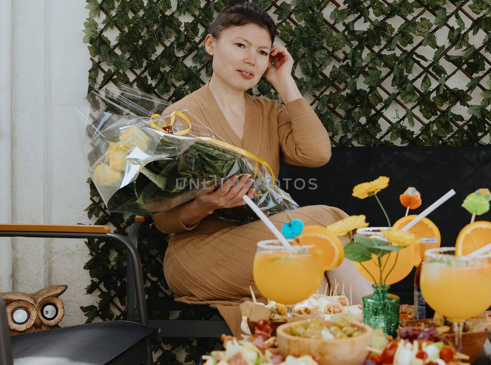 Portrait of one young beautiful Caucasian brunette girl in a knitted dress holding a bouquet of yellow roses in her hands, sitting on a garden chair in the backyard of a house on a spring sunny day and thoughtfully looking to the side, straightening her hair with her hand, celebrating her birthday, side view close-up.
