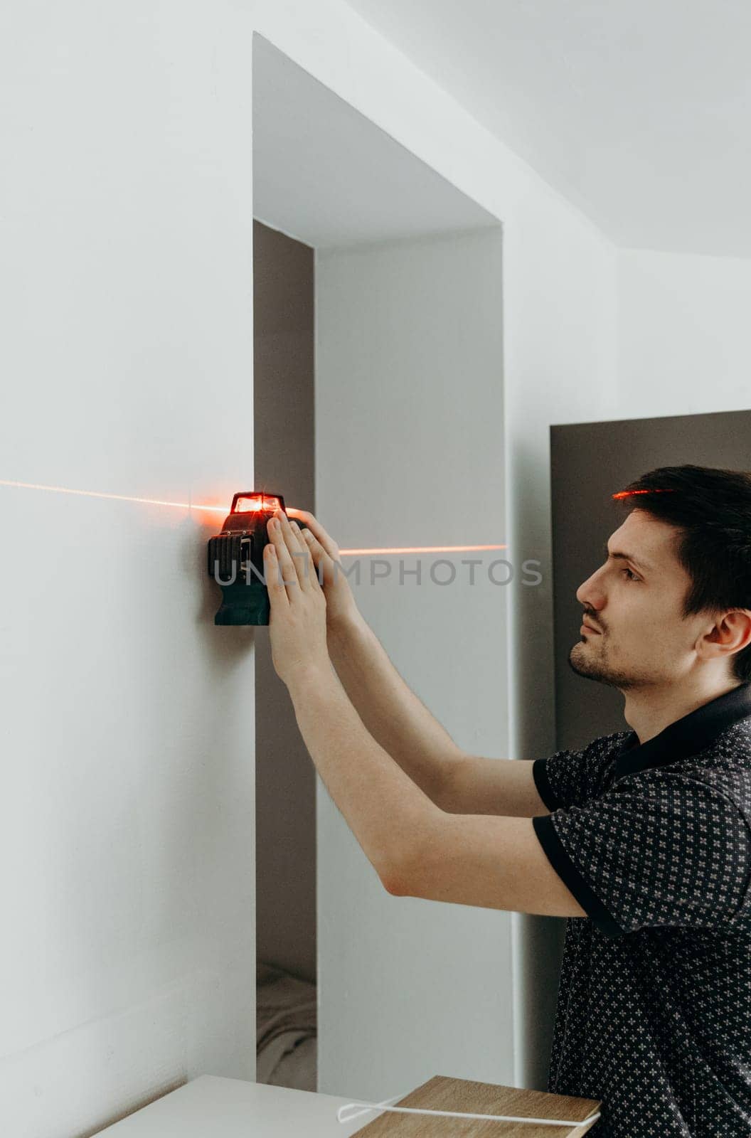 One young handsome brunette Caucasian guy stands sideways to the right and uses a modern laser level to measure a straight line on the wall before drilling with a drill, close-up side view.