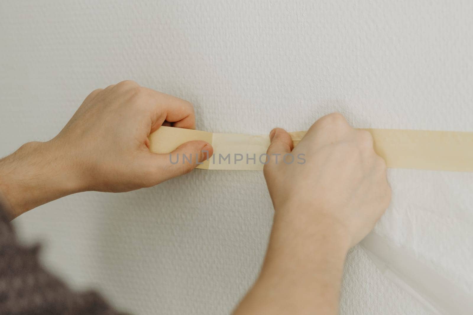 One young unrecognizable Caucasian guy glues with both hands adhesive tape with transparent cellophane on the wall to collect dust and debris, before drilling with a drill, side view close-up.