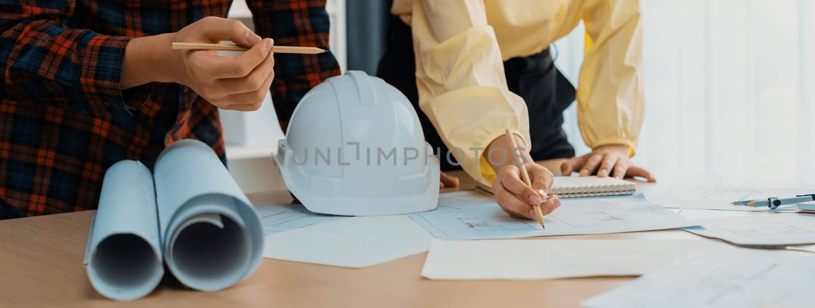 Professional engineer draws a blueprint on meeting table with blueprint and equipment scattered around while project manager using laptop analysis data at meeting room. Closeup. Delineation.