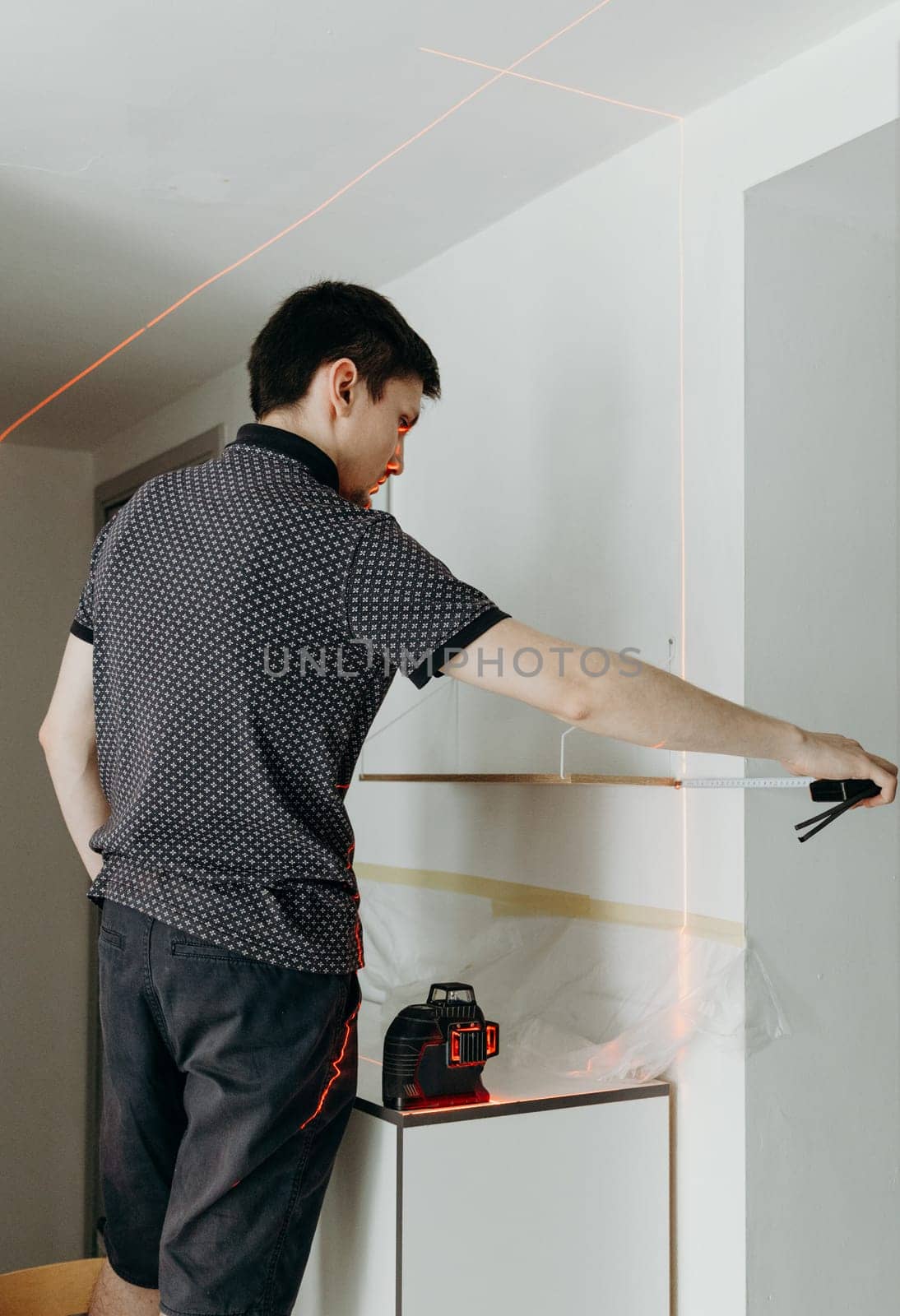 One young handsome Caucasian brunette guy stands half sideways, holds a measuring construction tape in his hands and checks the evenness of a hung wooden shelf on a white wall in a room on a spring day using a laser level, side view close-up with depth of field.