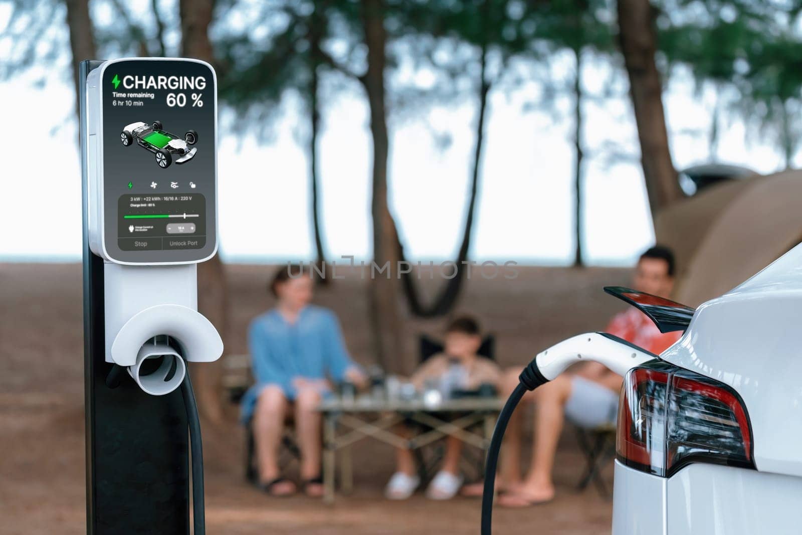 Alternative family camping trip traveling by the beach with electric car recharging battery from EV charging station with blurred family enjoying the seascape campsite background. Perpetual