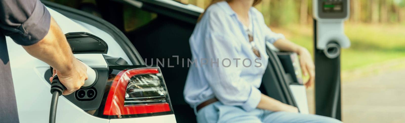 Panorama banner of lovely young couple recharging battery for electric car during autumnal road trip travel EV car in autumnal forest. Eco friendly travel on vacation during autumn. Exalt