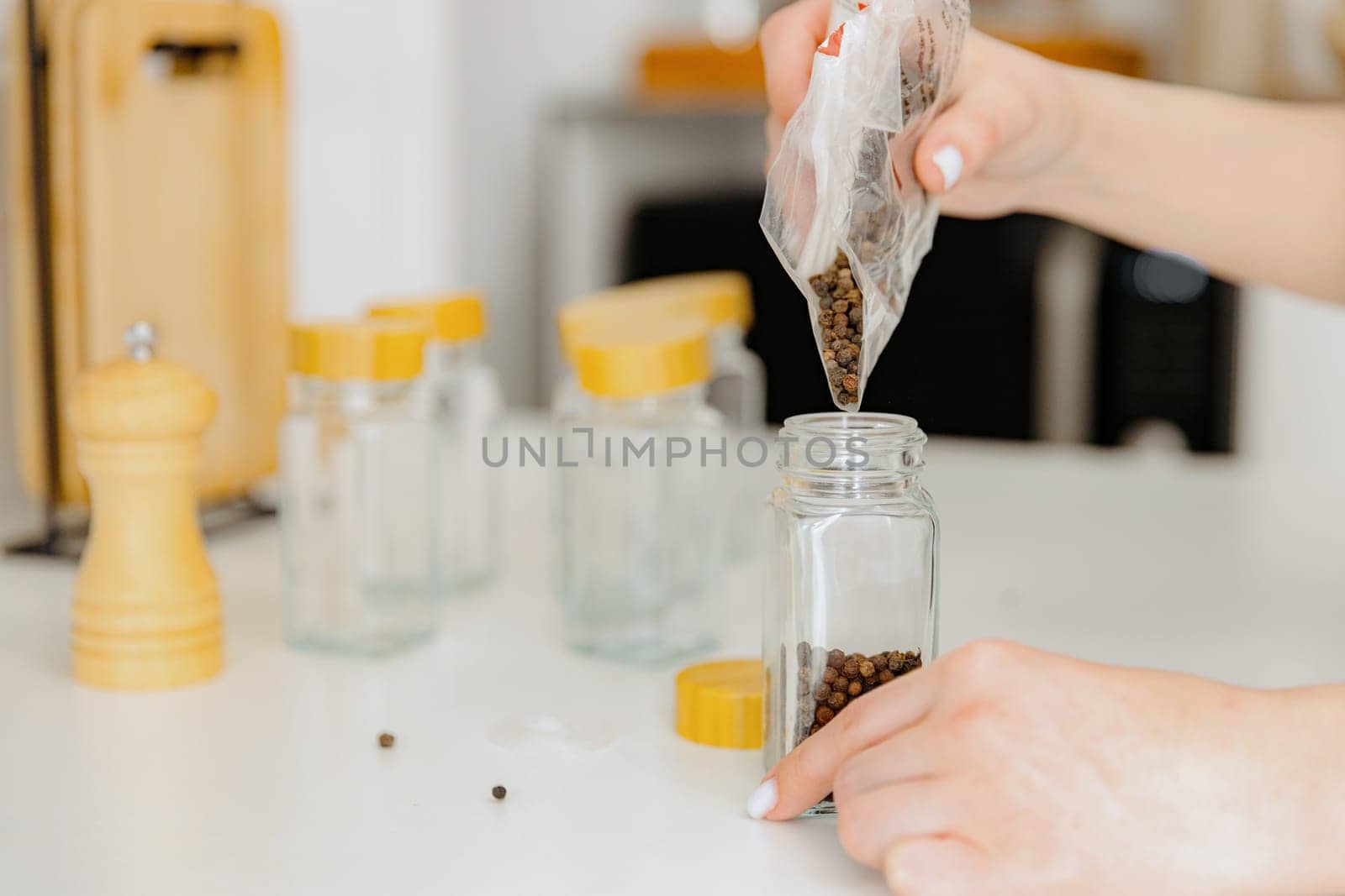 One young Caucasian unrecognizable girl pours seasoning spice black peppercorns from a plastic bag into a new glass transparent jar, standing at a white table in the kitchen on a summer day, side view close-up with depth of field. Eco-friendly storage concept.