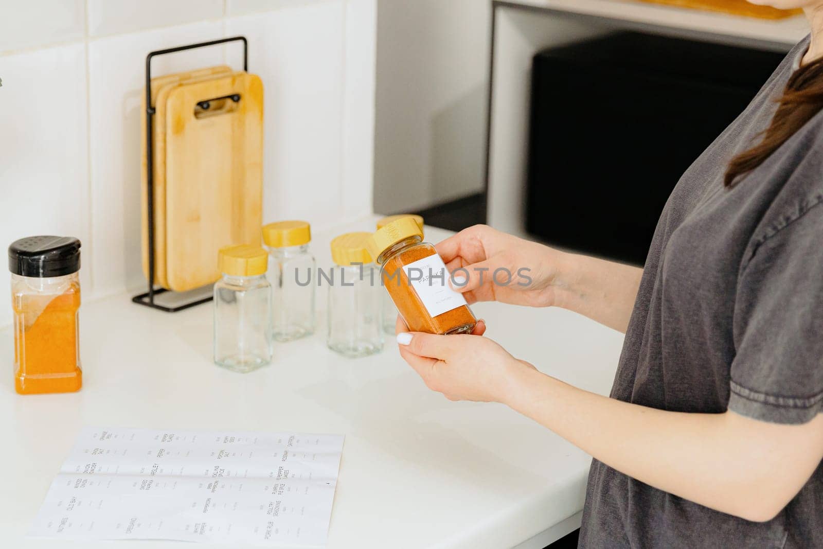 One young Caucasian unrecognizable girl holds a glass jar with a spice seasoning in her hands, having covered a sticker where it says paprika, standing at a white table in the kitchen on a summer day, side view close-up with depth of field. Eco-friendly storage concept.