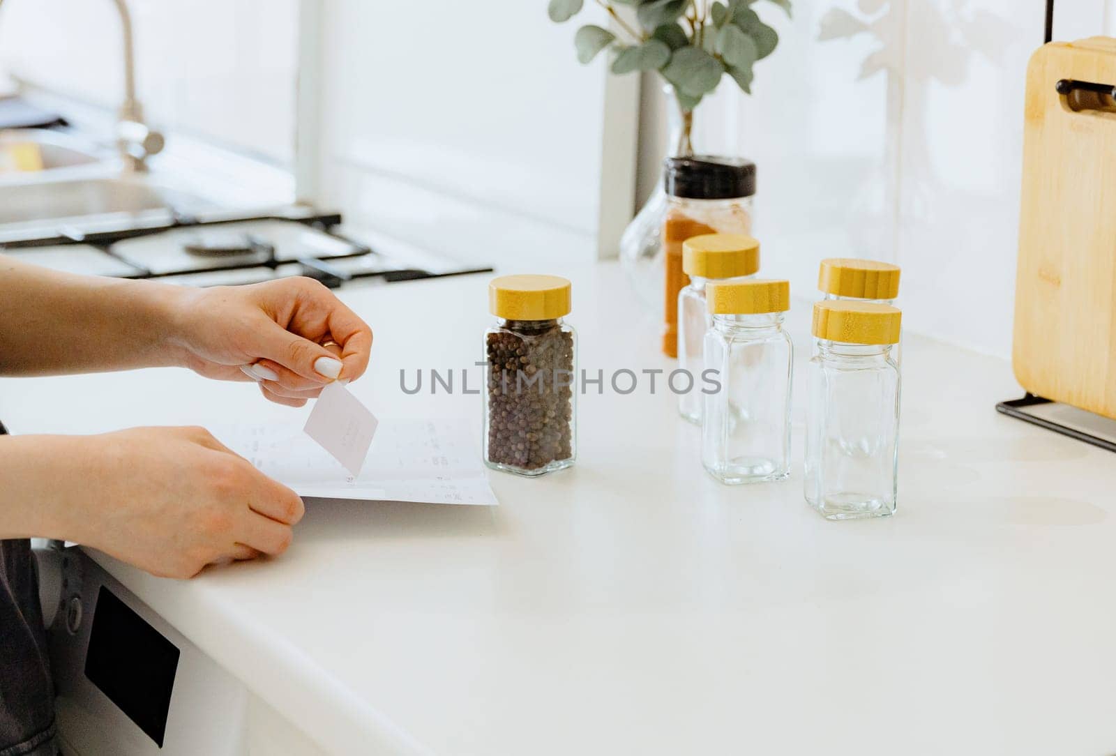 One young Caucasian unrecognizable girl peels off a sticker for a glass jar with seasoning spice black peppercorns, standing at a white table in the kitchen on a summer day, side view close-up with depth of field. Eco-friendly storage concept.