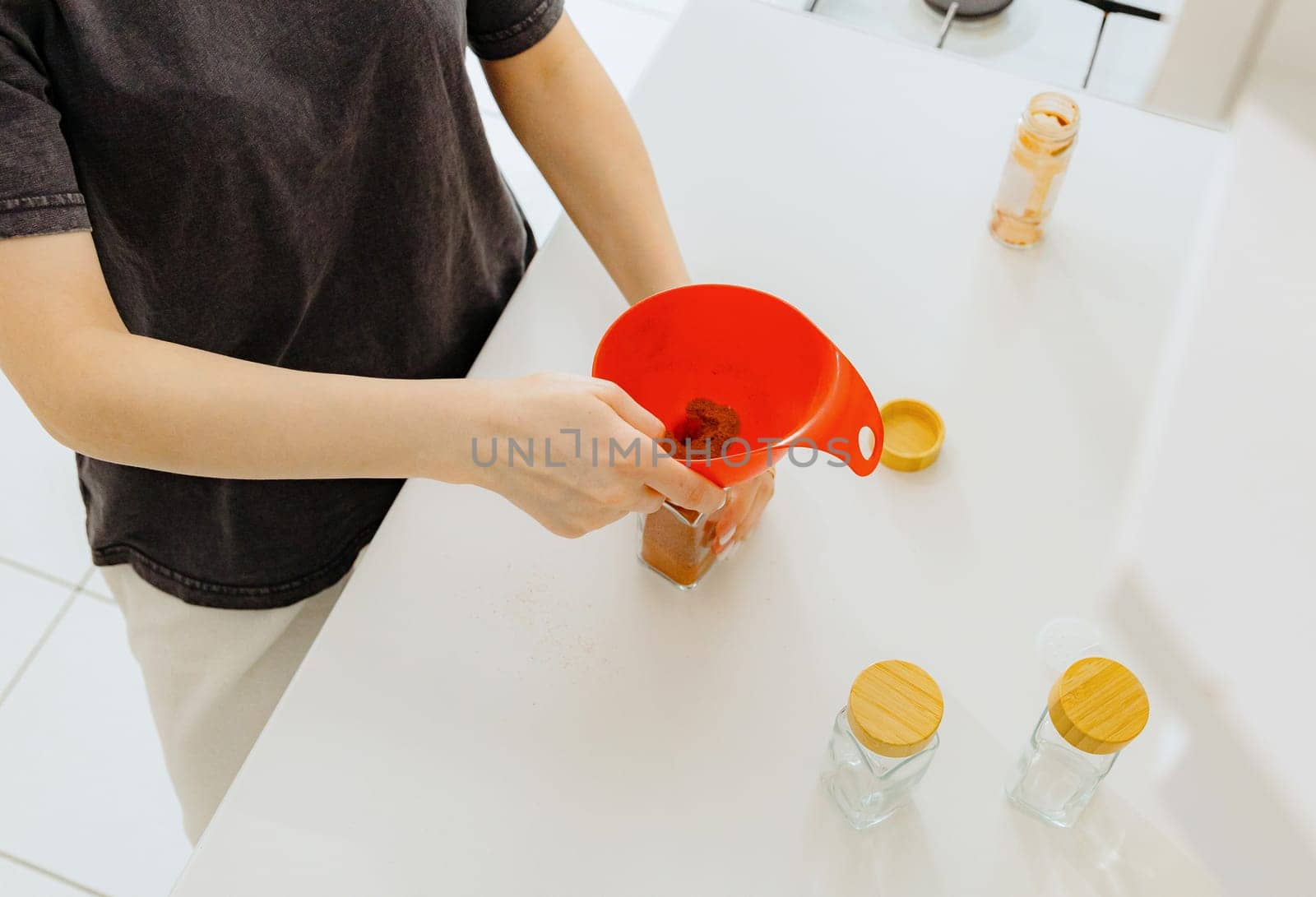 One young Caucasian unrecognizable girl pours seasoning spice ground paprika into a new glass transparent jar, standing at a white table in the kitchen on a summer day, using a red watering can, top view close-up with depth of field. Eco-friendly storage concept.