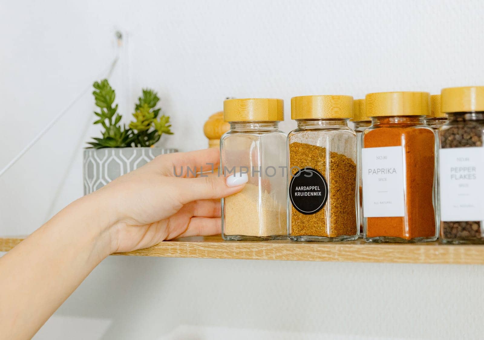 One young Caucasian unrecognizable girl takes out with one hand a glass jar with the spice dry ground garlic from a wooden wall shelf in the kitchen on a summer day, side view close-up with depth of field. Eco-friendly storage concept.