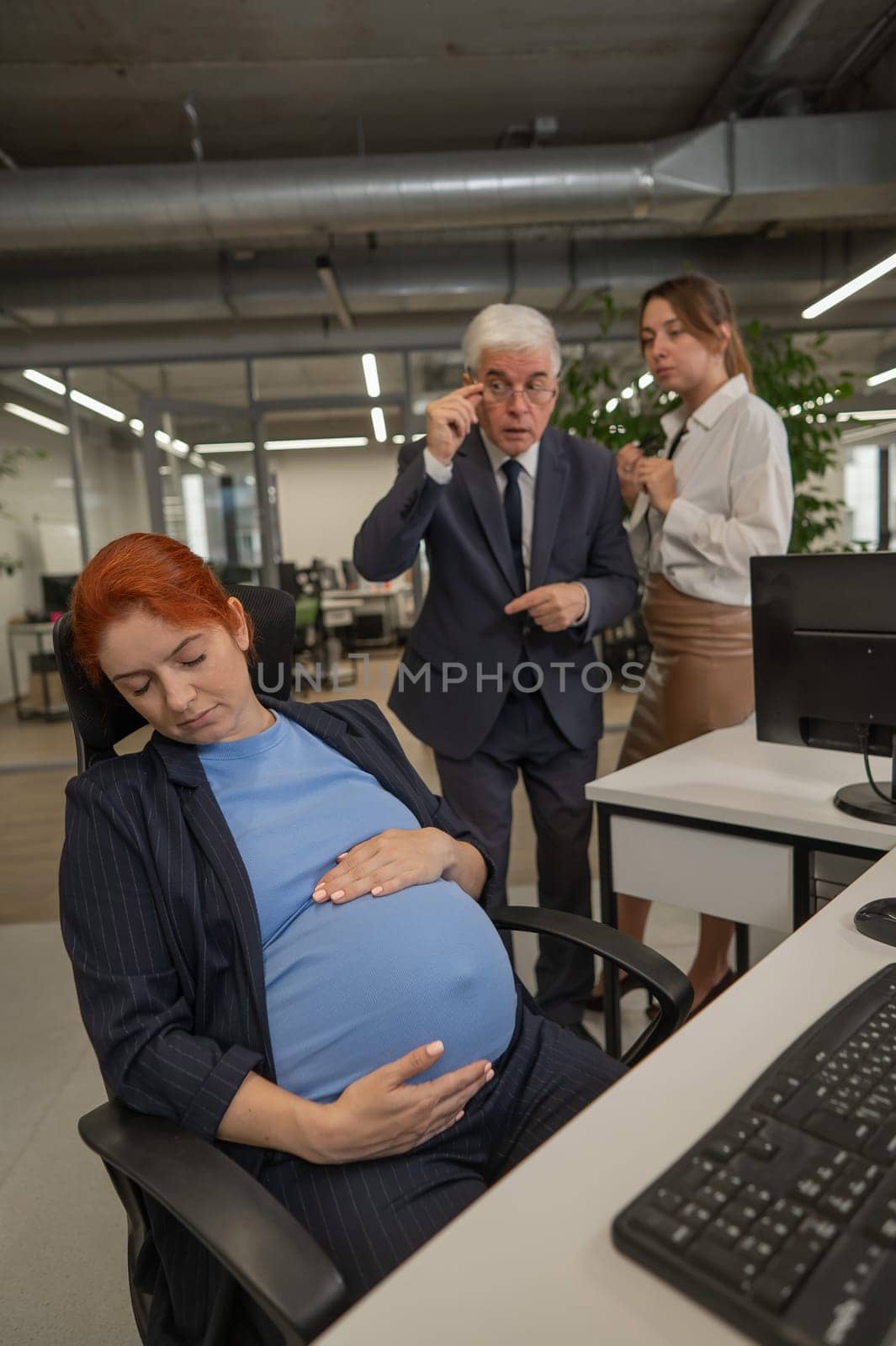 Elderly Caucasian man and woman gossiping behind their sleeping pregnant colleague in the office. Vertical photo