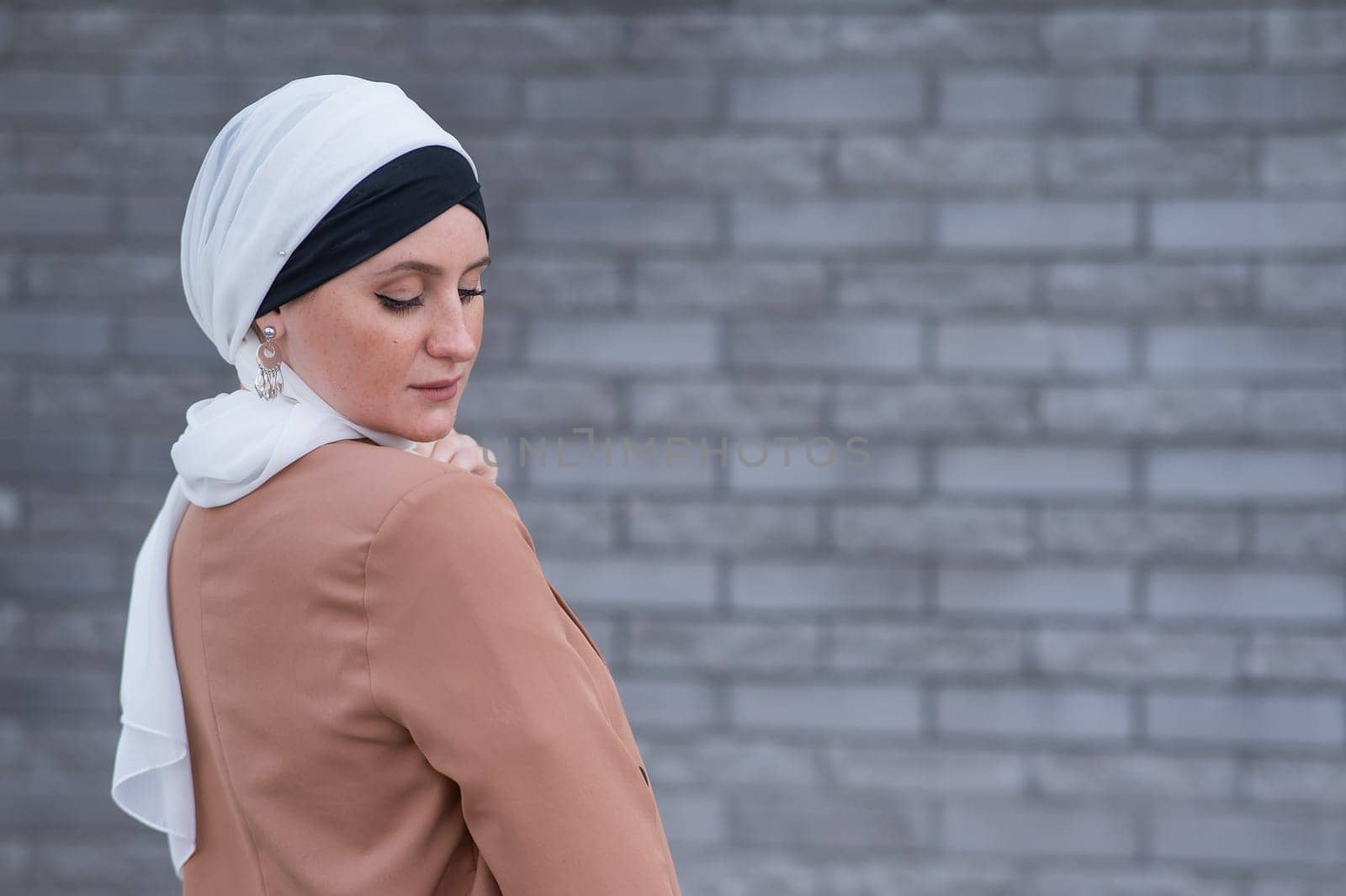 Portrait of a young blue-eyed woman in a hijab against a gray brick wall. A Muslim woman looks at the camera turning around. by mrwed54
