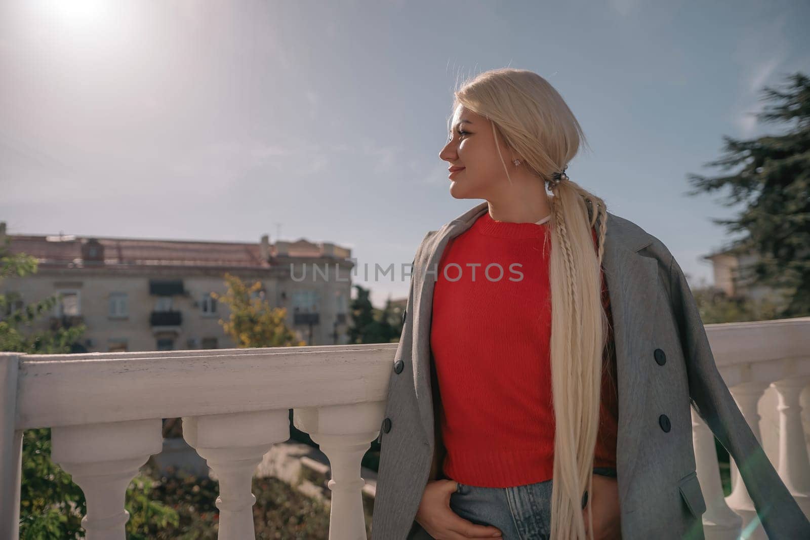 A blonde woman wearing a red shirt and gray jacket stands on a balcony. She is wearing ripped jeans and has her hands in her pockets