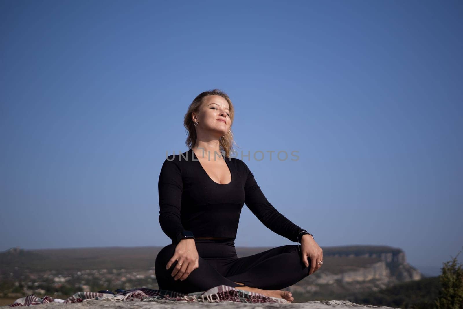 A woman is sitting on a rock with her legs crossed and her hands on her knees. She is looking up at the sky and she is in a peaceful and relaxed state