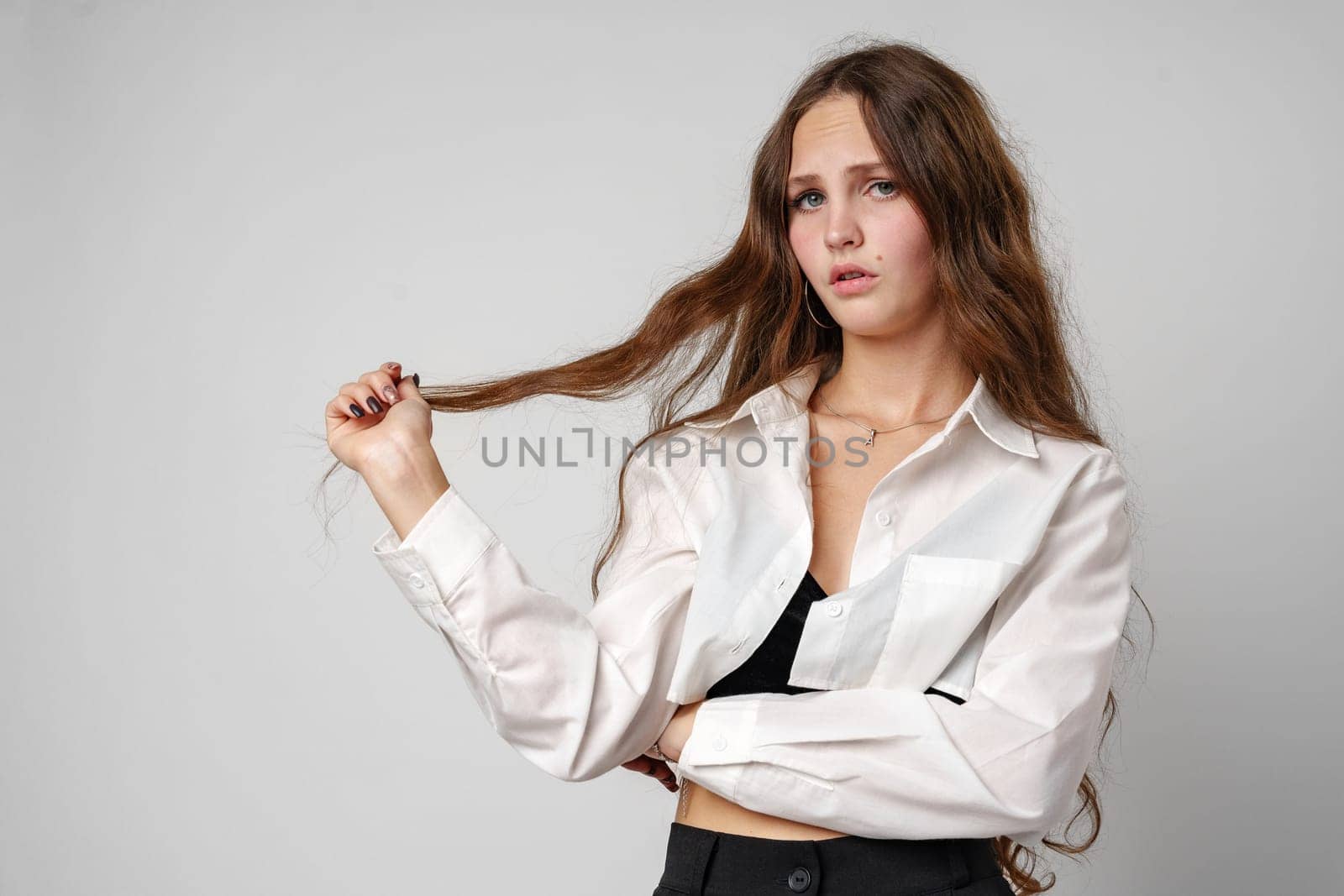 A young woman stands against a plain background, her gaze fixed off-camera with a serious expression. She is wearing a white blouse and a black tank top, holding a portion of her long brown hair in her hand and appears to be either contemplating something or posing pensively.