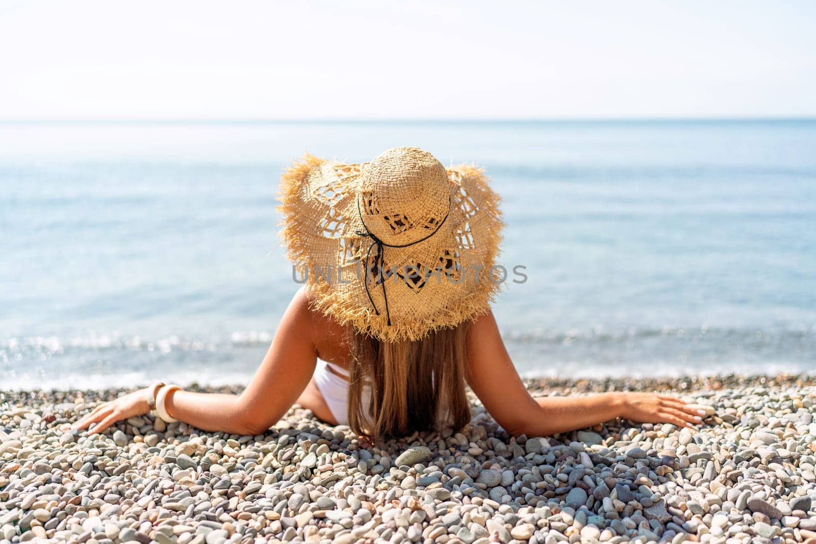 Beach Relaxation woman in hat sits on a pebble beach enjoying the sunshine. The concept of travel, vacation at sea.