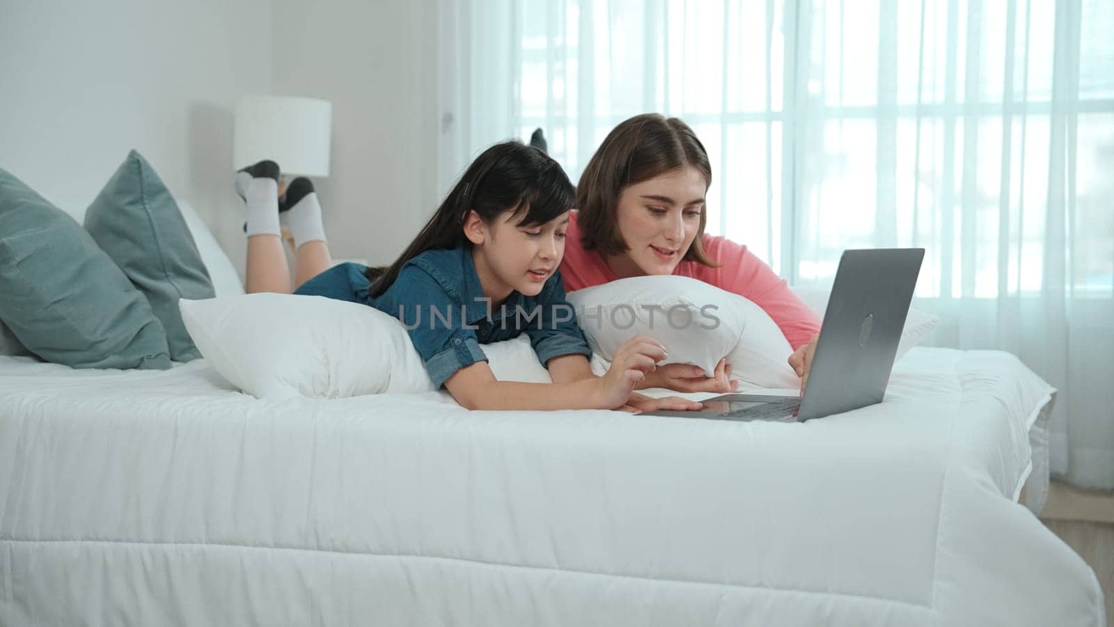 Attractive daughter and caucasian mother watching movie at laptop screen while lie on bad. Happy family spend time together before bed time. Parent and cute girl enjoy playing games together Pedagogy.