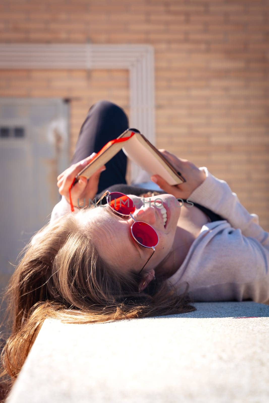 Young woman holding book up in air, low angle view looking at camera outdoors by mariaphoto3