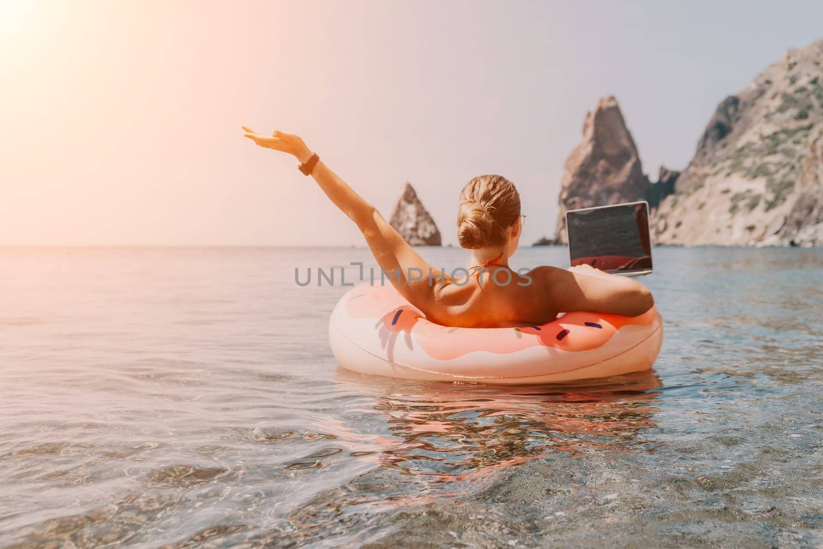 Woman freelancer works on laptop swimming in sea on pink inflatable ring. Pretty lady typing on computer while floating in the sea on inflatable donut at sunset. Freelance, remote work on vacation