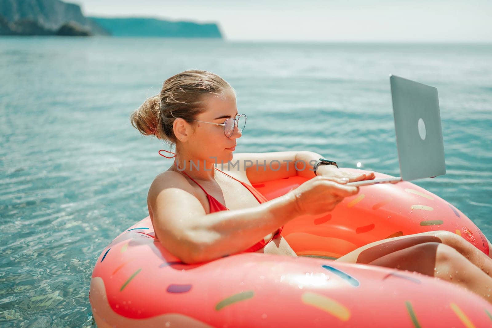 Woman laptop sea. Freelancer woman in sunglases floating on an inflatable big pink donut with a laptop in the sea. People summer vacation rest lifestyle concept