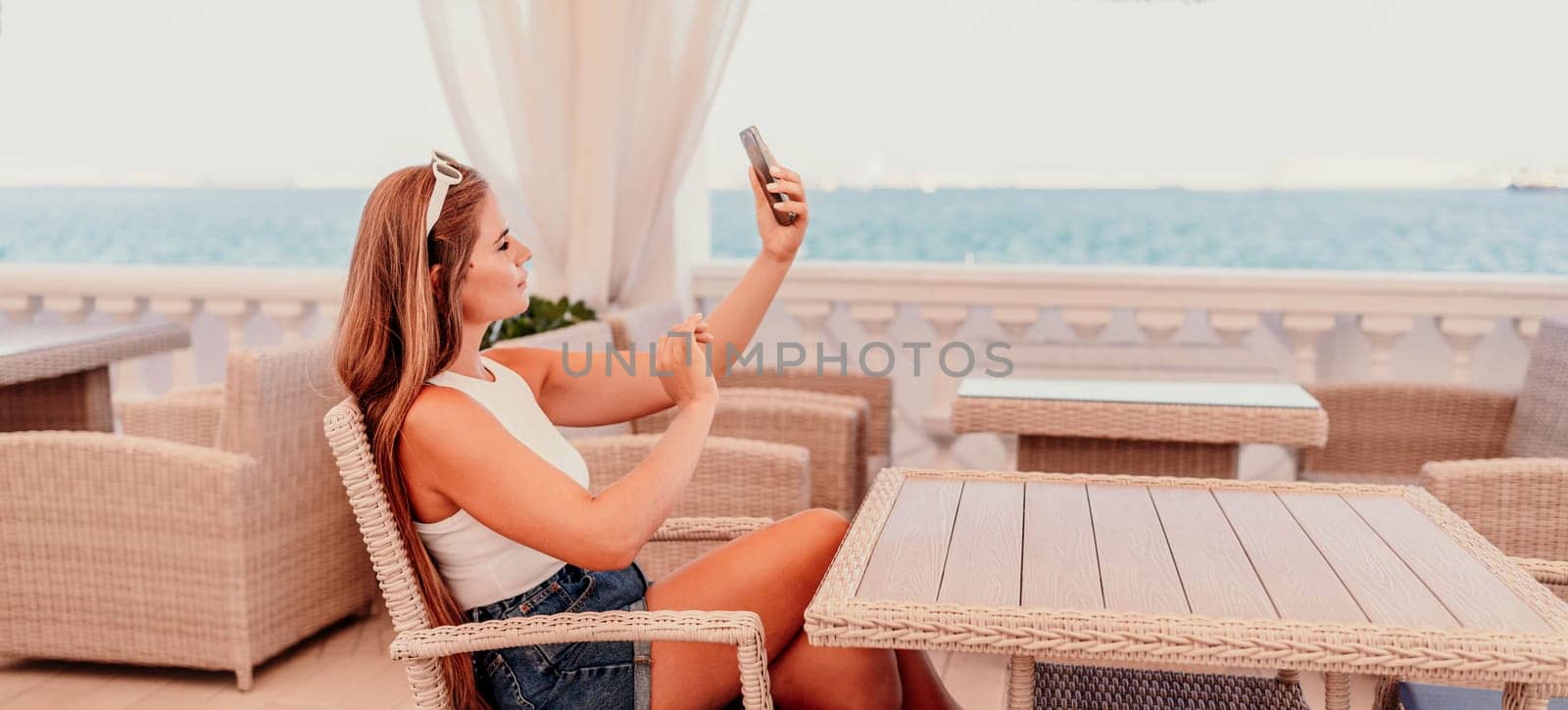 A woman is sitting at a table with a cell phone in her hand. She is taking a picture of the ocean