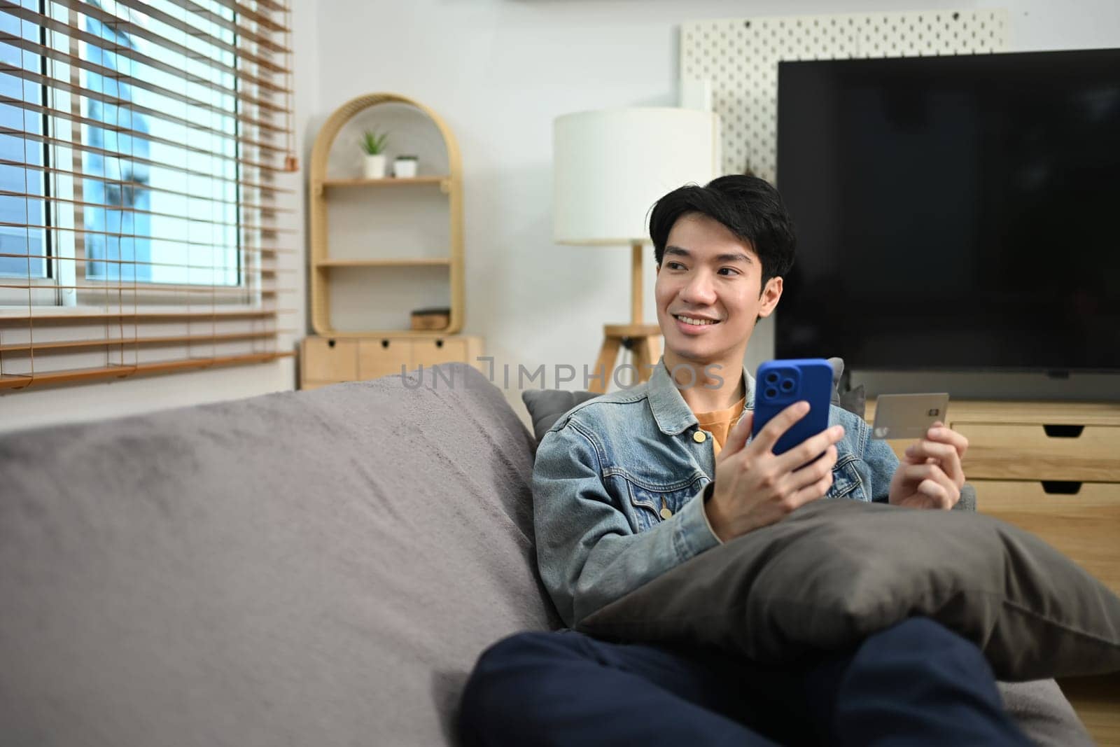 Smiling young man with mobile phone and credit card looking away while sitting on sofa.