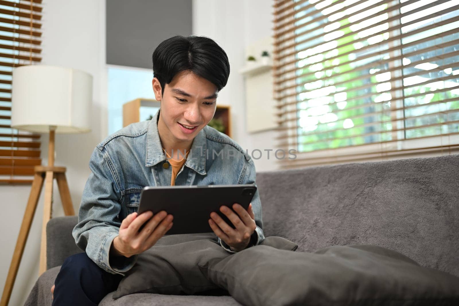 Smiling asian male freelancer sitting on couch and working with digital tablet.