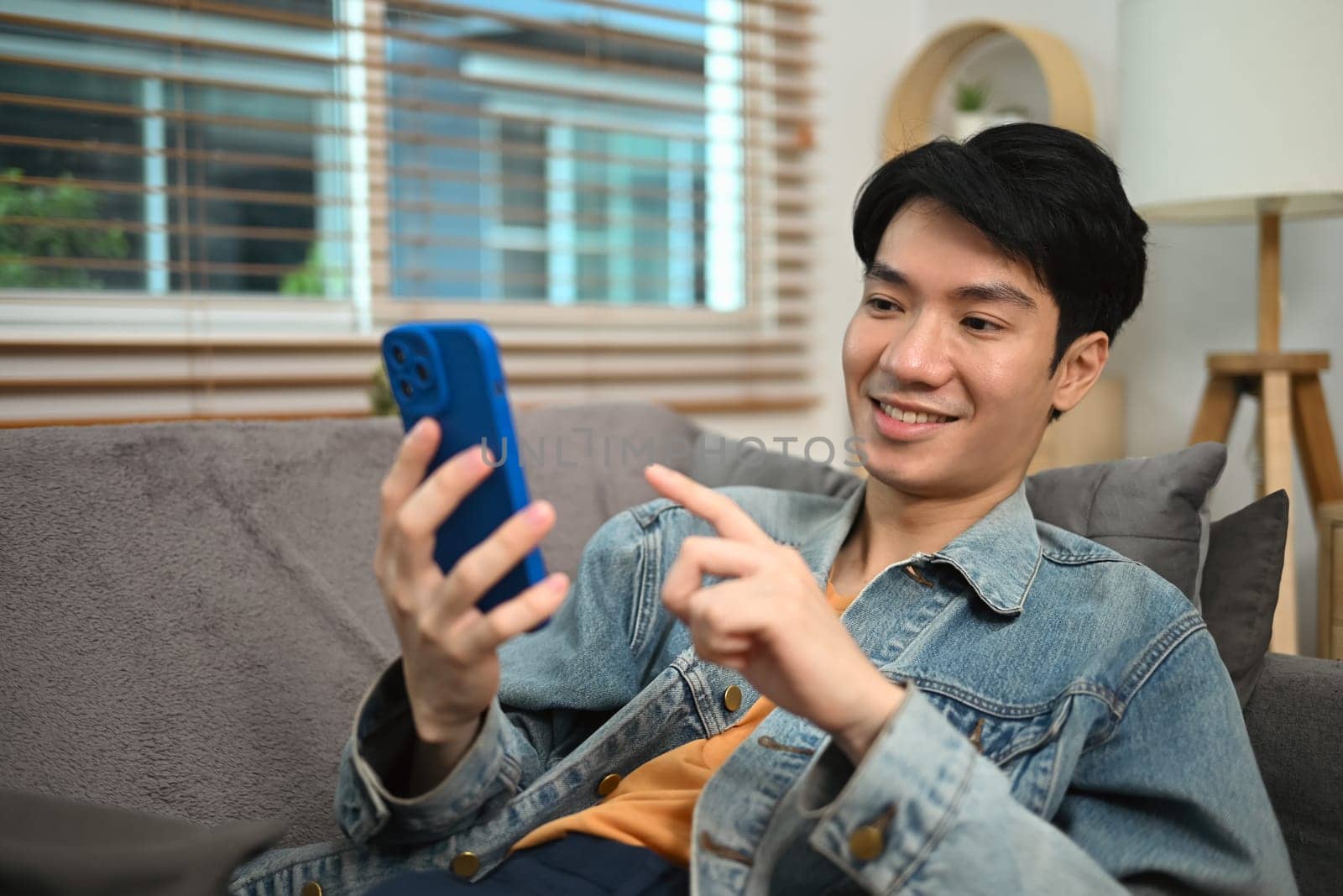 Cheerful young man typing text message on mobile phone while relaxing in living room.