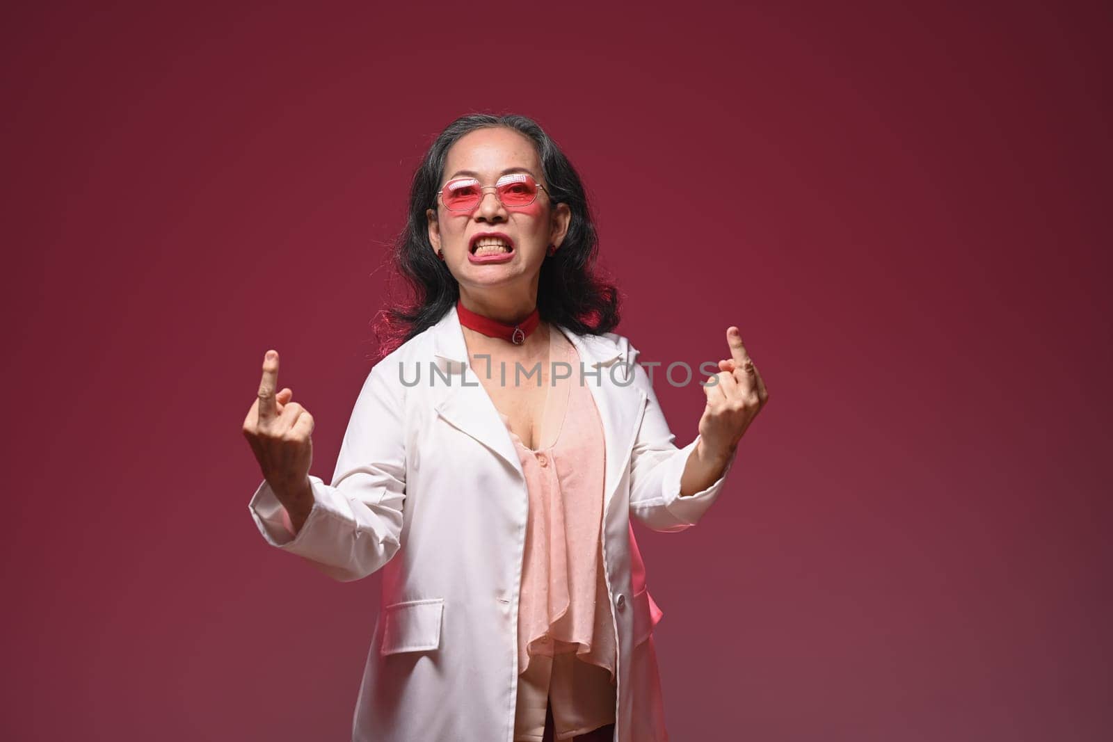 Studio shot of crazy funny mature woman showing middle finger gesture over red background.
