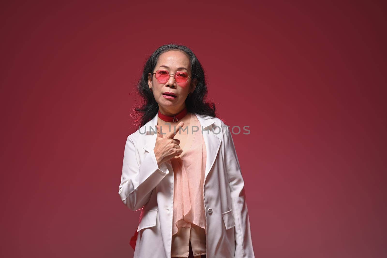 Cool and stylish middle age woman wearing pink glasses and fashionable clothes on red background.