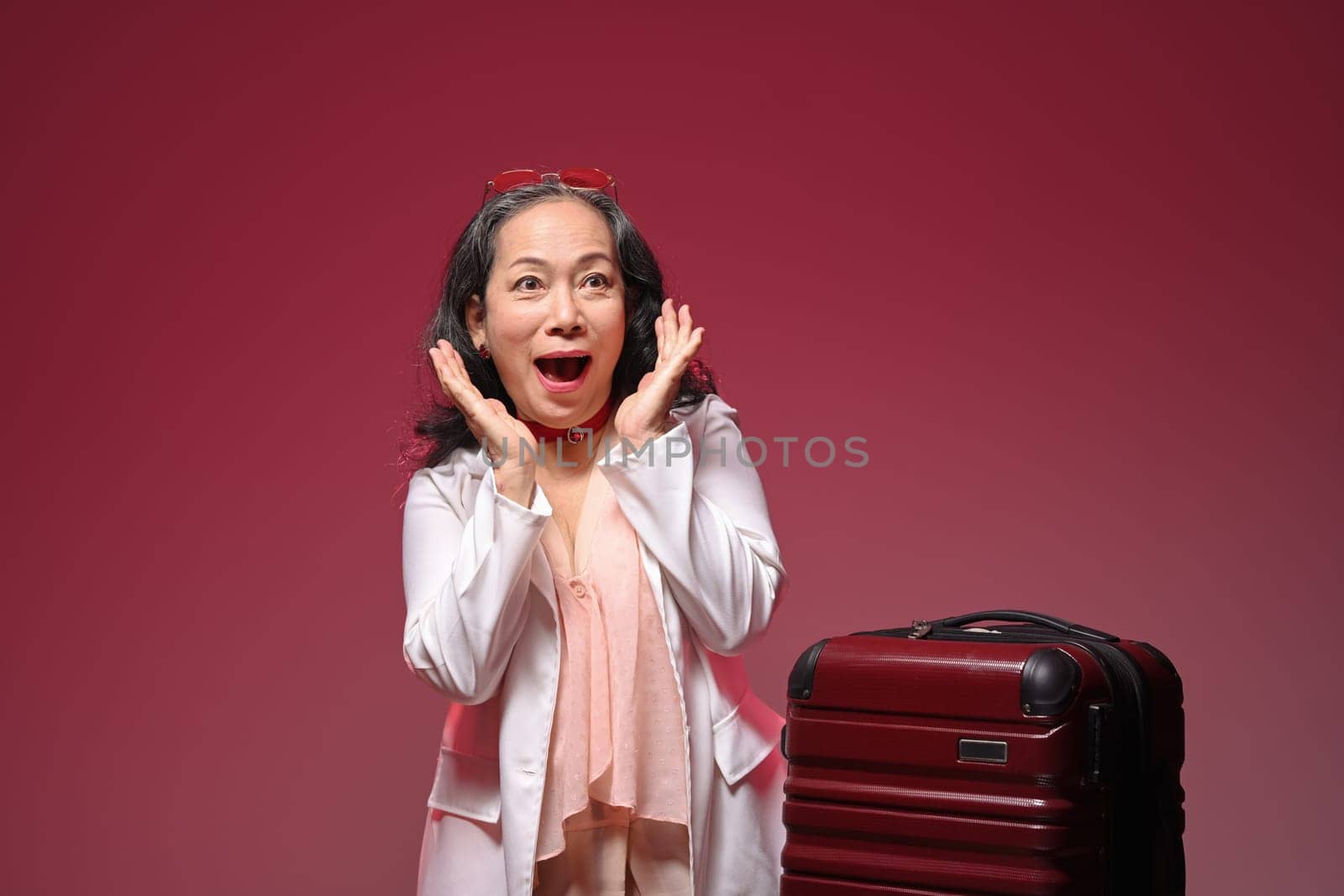 Happy and excited senior woman with suitcase over red background. Summer, travel and vacation concept.