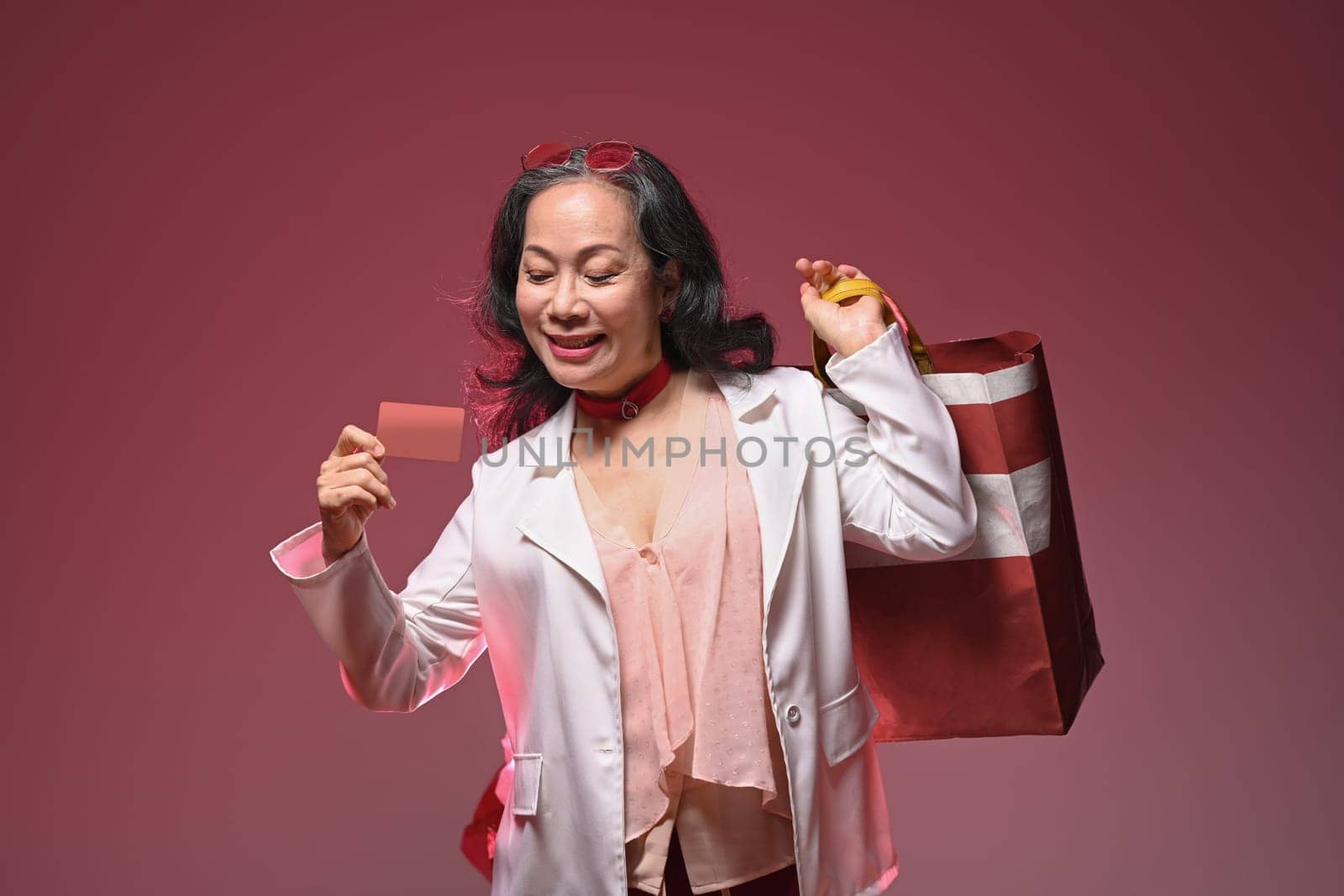 Stylish middle aged woman holding credit card and shopping bags over red background.