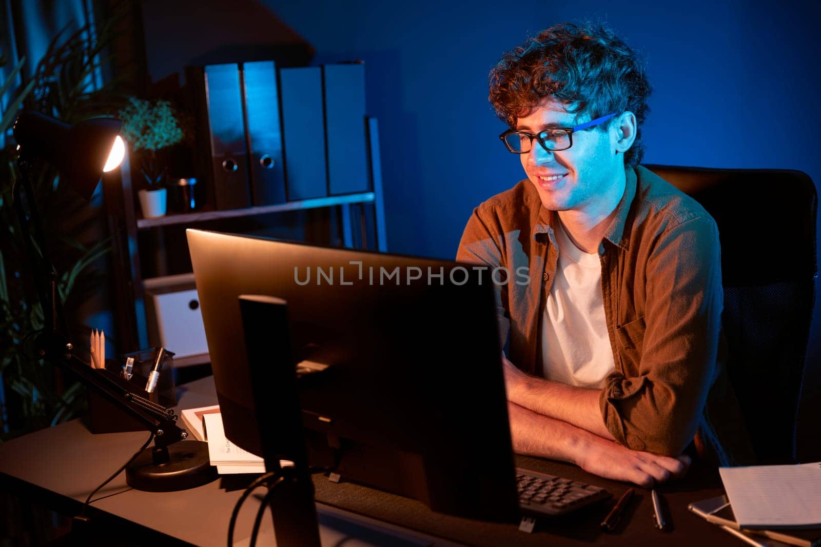 Smiling young businessman focusing on pc for creative startup project job at neon light modern office. Thinking marketing network online planning of creator to market channel design concept. Gusher.