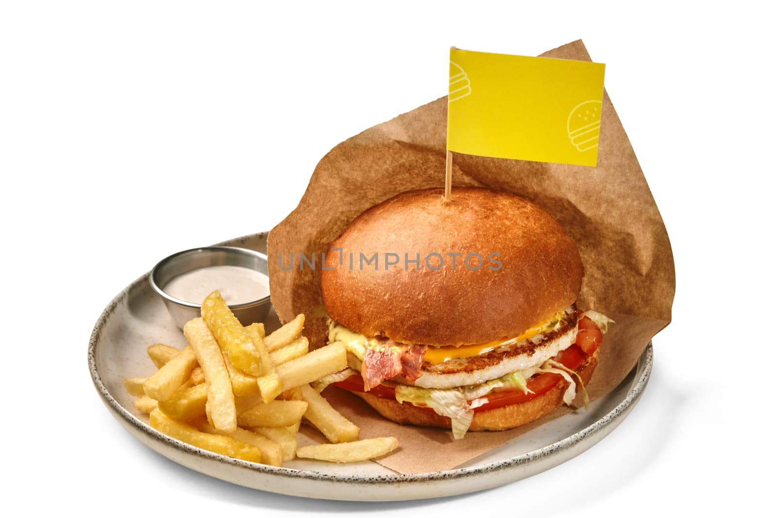 Appetizing burger, marked by yellow Chicken Burger flag, with juicy patty, fried bacon, vegetables and cheese served with crispy fries and dipping sauce on ceramic plate, against white background